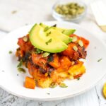 Mayan harvest bake on a white plate with layers of polenta and quinoa with roasted sweet potatoes, plantains. Topped with avocado.