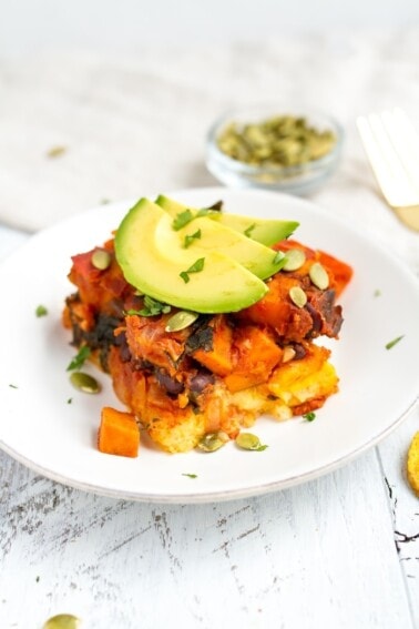 Mayan harvest bake on a white plate with layers of polenta and quinoa with roasted sweet potatoes, plantains. Topped with avocado.
