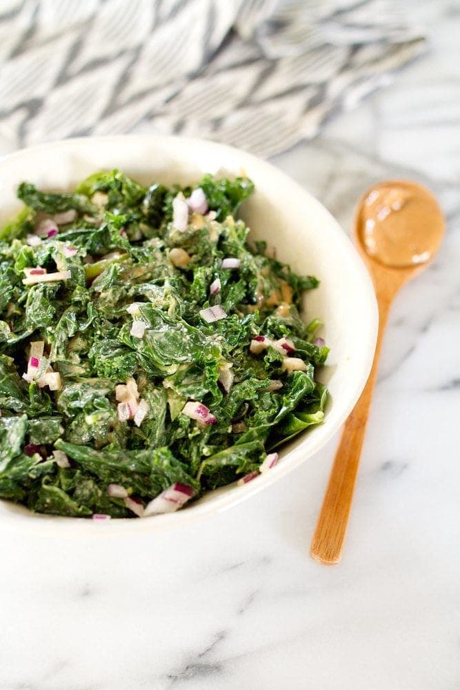 A bowl of kale salad with spicy peanut dressing with a wooden spoon laying next to it.