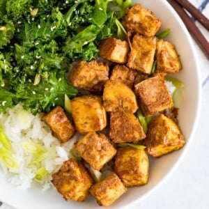 White bowl with kale topped with sesame seeds, crispy baked peanut tofu, and rice topped with scallions. Chop sticks are next to the bowl. The bowl is sitting on a blue and white striped folded napkin.