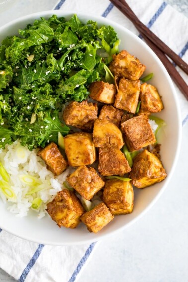 White bowl with kale topped with sesame seeds, crispy baked peanut tofu, and rice topped with scallions. Chop sticks are next to the bowl. The bowl is sitting on a blue and white striped folded napkin.