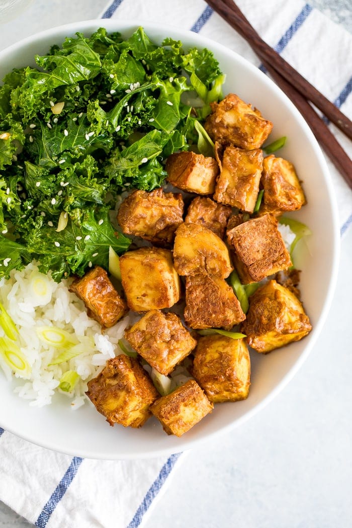 Bowl with kale topped with sesame seeds, crispy baked peanut tofu, and rice topped with scallions.