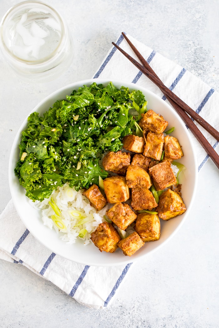 Bowl with kale topped with sesame seeds, crispy baked peanut tofu, and rice topped with scallions. A glass of water and chop sticks are next to the bowl.
