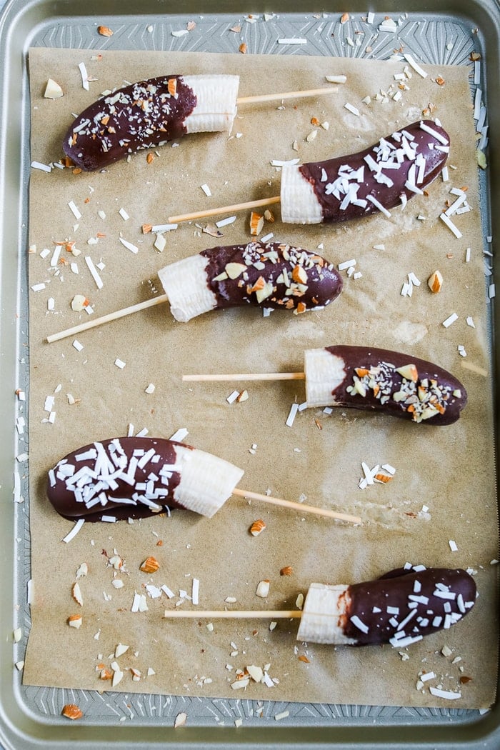 Chocolate covered banana pops on parchment paper lined cookie sheet and sprinkled with almonds and coconut.