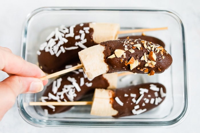 Hand holding a frozen banana on a stick dipped in chocolate and sprinkled with almonds. Glass container with chocolate covered bananas sprinkled with coconut.