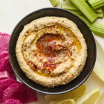 Creamy hummus with olive and spices on top in a black serving bowl with celery and chopped veggies displayed around the bowl.