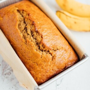 Baked whole wheat banana bread in a loaf pan with bananas in the background.