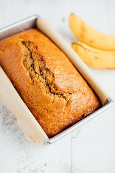 Baked whole wheat banana bread in a loaf pan with bananas in the background.