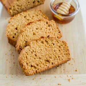 Three slices of honey whole wheat brown bread with the remaining loaf on a cutting board. There is a small dish of honey with a wooden honey comb resting inside the dish.