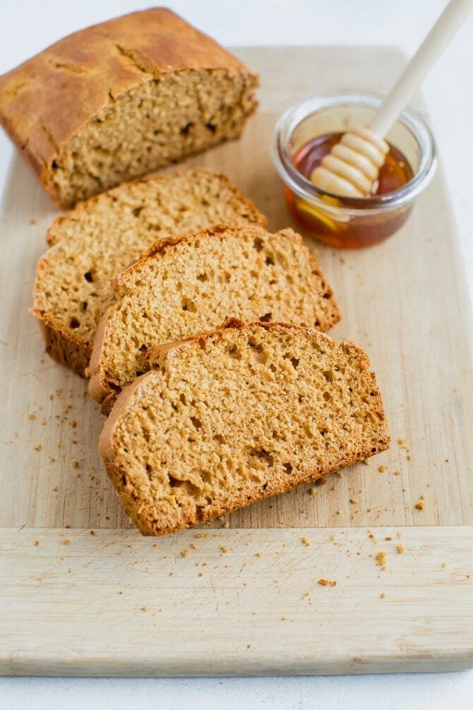 Three slices of honey whole wheat brown bread with the remaining loaf on a cutting board. There is a small dish of honey with a wooden honey comb resting inside the dish.