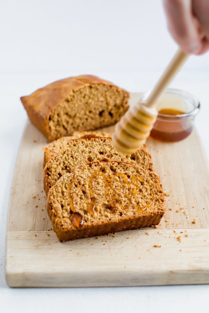 A wooden honey comb is drizzling honey onto a slice of whole wheat honey brown bread. The loaf of bread is in the background. 