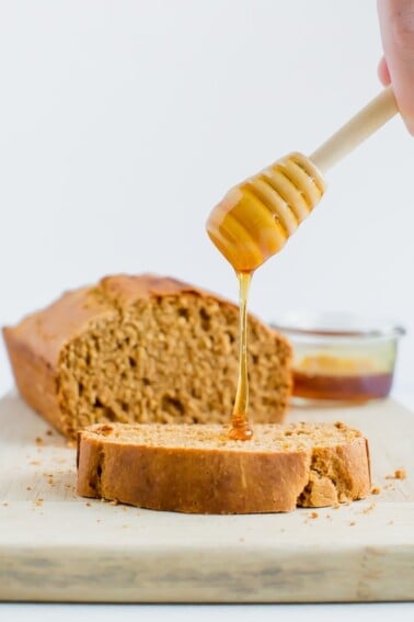 A wooden honey comb is drizzling honey onto a slice of whole wheat honey brown bread. The loaf of bread is in the background.