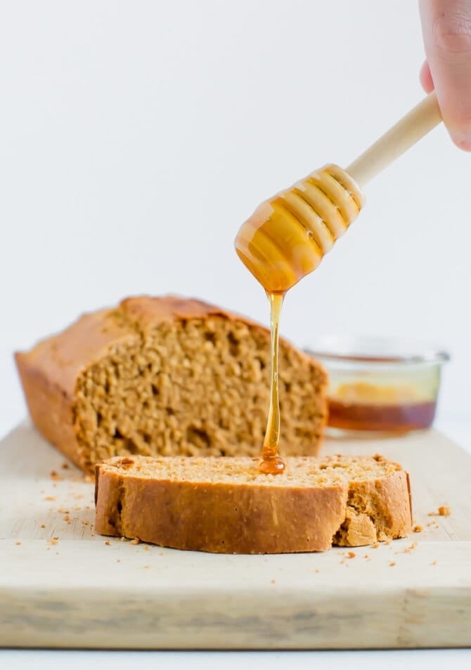 A wooden honey comb is drizzling honey onto a slice of whole wheat honey brown bread. The loaf of bread is in the background. 