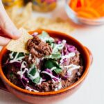 Ceramic bowl of black bean dip, garnished with cilantro and purple onion. A persons hand is holding a tortilla chip with bean dip in front of the bowl. A smaller bowl of round carrot slices, cans of La Croix and some tortilla chips are in the background.