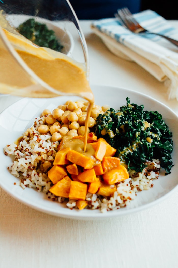 White bowl with brown rice, roasted sweet potato, chickpeas, kale and a peanut sauce being poured from a measuring cup.