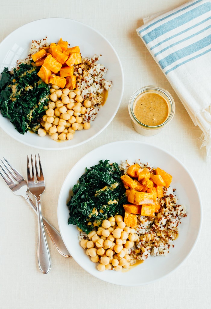 Two white bowls with brown rice, roasted sweet potato, chickpeas and kale. Drizzled with peanut sauce. The jar of sauce is off to the side.