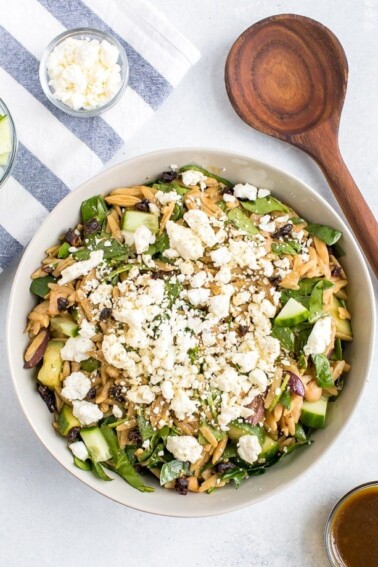 Bowl of orzo pasta salad with feta, olives, spinach, cucumber and white beans. Wood spoon and bowls of cucumber and feta on a dish towel next to the bowl.