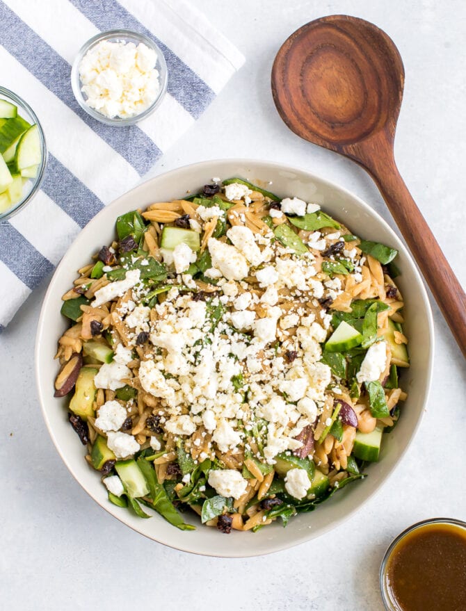 Bowl of orzo pasta salad with feta, olives, spinach, cucumber and white beans. Wood spoon and bowls of cucumber and feta on a dish towel next to the bowl.