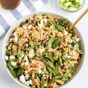 Orzo pasta salad tossed with a balsamic dressing. Orzo mixed with spinach, cucumber, feta, sun-dried tomatoes, olives and seasoning. Bowls of cucumber, feta and balsamic dressing in the background.