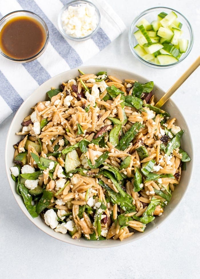 Orzo pasta salad tossed with a balsamic dressing. Orzo mixed with spinach, cucumber, feta, sun-dried tomatoes, olives and seasoning. Bowls of cucumber, feta and balsamic dressing in the background.