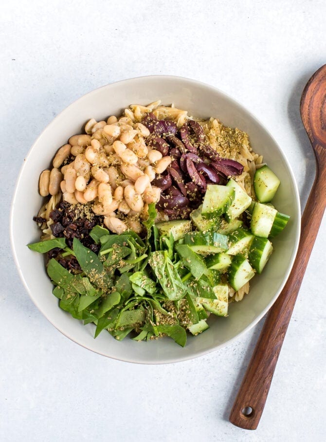 Ingredients for orzo pasta salad in a bowl. White beans, sun dried tomato, kalamata olives, cucumber, spinach and seasoning.