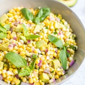 Bowl of avocado and corn salad topped with fresh basil leaves. Cut open avocado in the background.