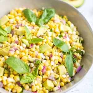 Bowl of avocado and corn salad topped with fresh basil leaves. Cut open avocado in the background.