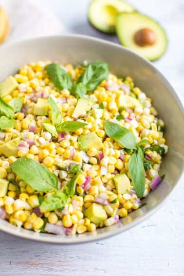 Bowl of avocado and corn salad topped with fresh basil leaves. Cut open avocado in the background.