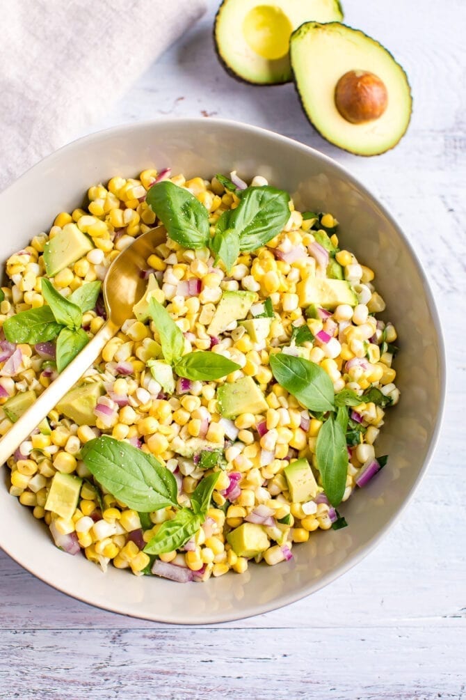 Bowl of raw corn and avocado salad mixed with basil and red onion. Avocado halves and a napkin are beside the bowl. Spoon is in the bowl.