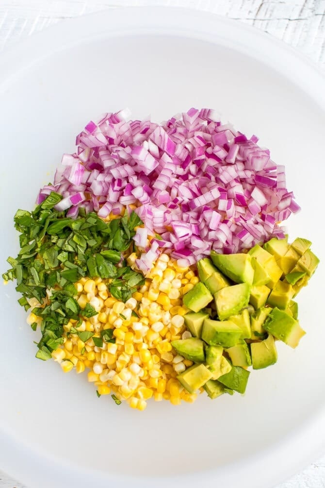 Ingredients for corn and avocado salad in a bowl including basil, corn, red onion and avocado.