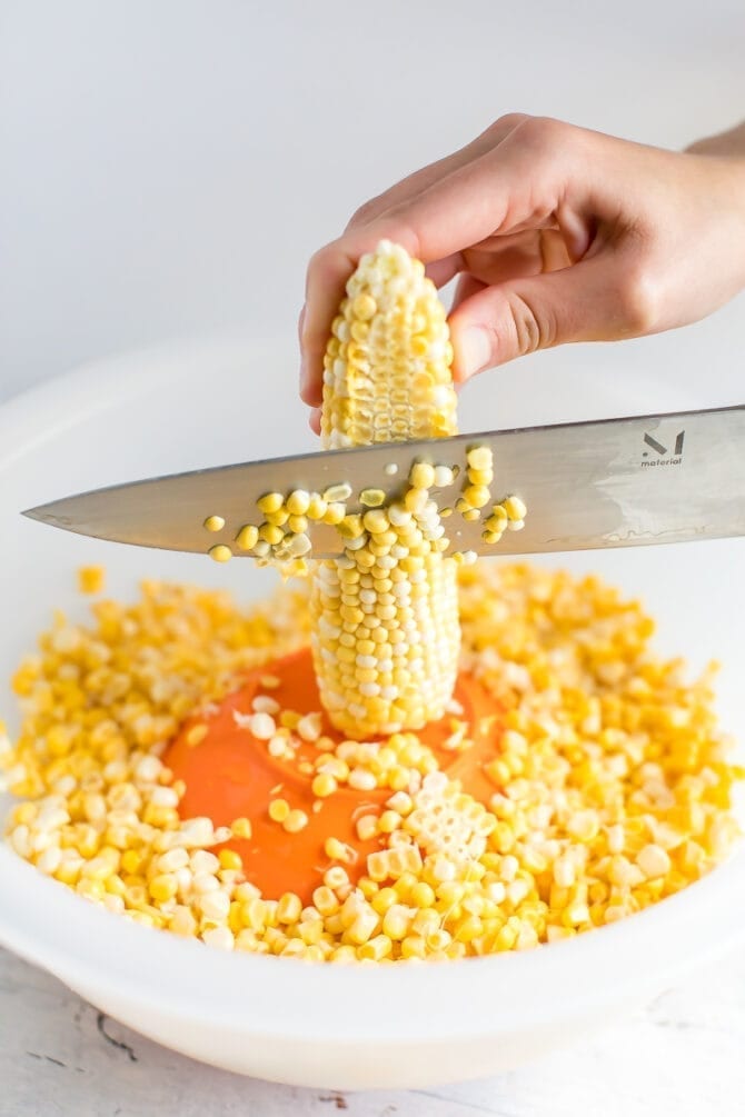 Hands cutting corn off of the cob. The corn cob is propped on a small bowl upside down in a larger bowl.