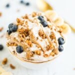 A bowl of yogurt covered with peanut butter drizzle, grain-free granola, blueberries and bananas in a gold rimmed bowl on a white wooden board. . A gold spoon in the background.