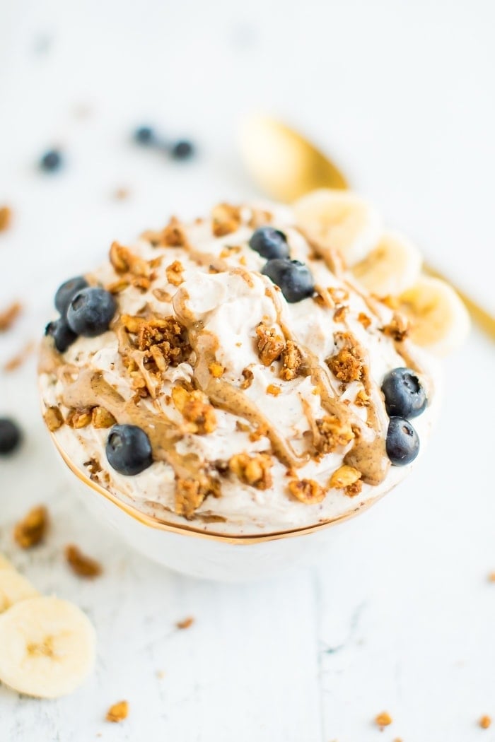 A bowl of yogurt covered with peanut butter drizzle, grain-free granola, blueberries and bananas in a gold rimmed bowl on a white wooden board. . A gold spoon in the background.
