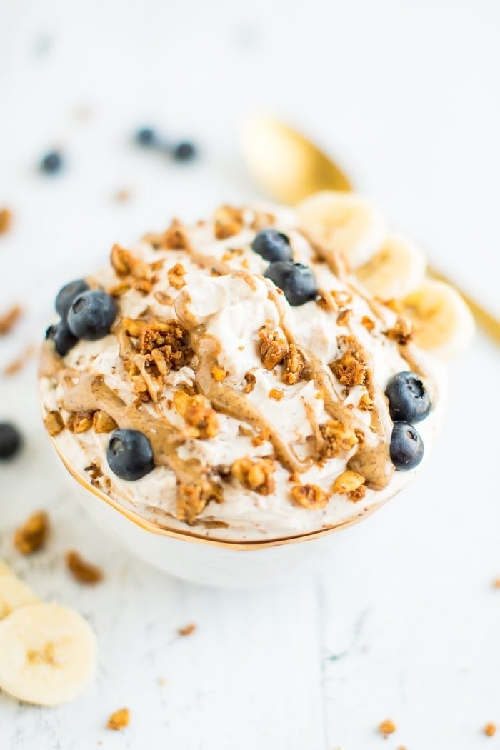 A delicious yogurt bowl made with peanut butter, grain-free granola, blueberries and bananas in a gold rimmed bowl with a gold spoon on a white wooden board.