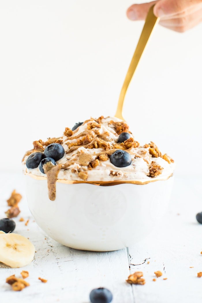 A bowl of yogurt covered with peanut butter drizzle, grain-free granola, blueberries and bananas in a gold rimmed bowl on a white wooden board. A hand is holding a gold spoon scooping the contents in the bowl.