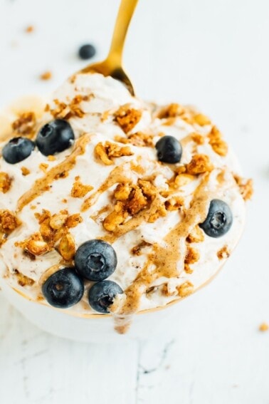 A bowl of yogurt covered with peanut butter drizzle, grain-free granola, blueberries and bananas in a gold rimmed bowl on a white wooden board. . A gold spoon is in the bowl.