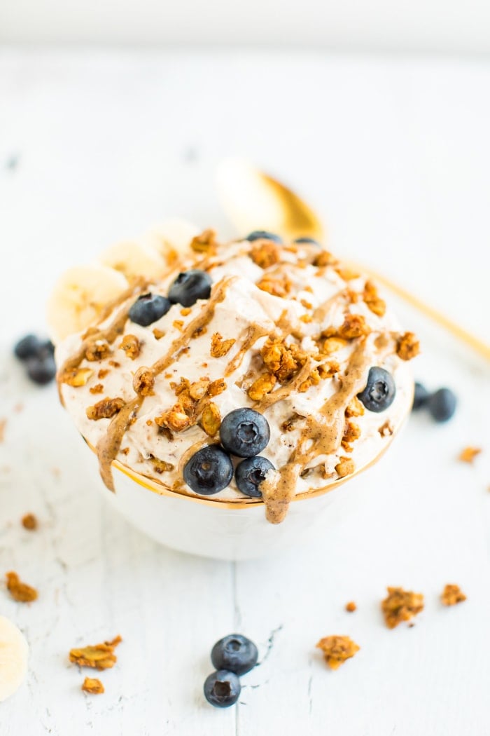 A bowl of yogurt covered with peanut butter drizzle, grain-free granola, blueberries and bananas in a gold rimmed bowl on a white wooden board. . A gold spoon in the background.