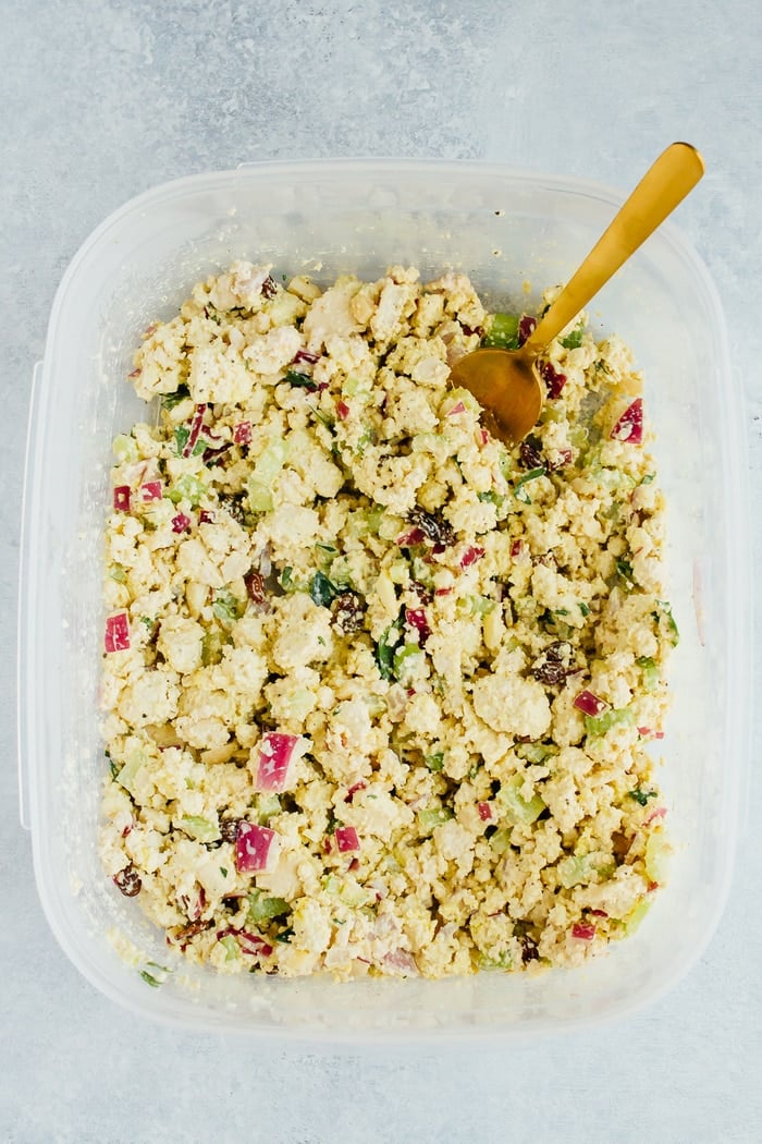 Overhead shot of clear tupperware of Vegan Chicken Salad made with tofu, celery, almonds and raisins on white countertop.