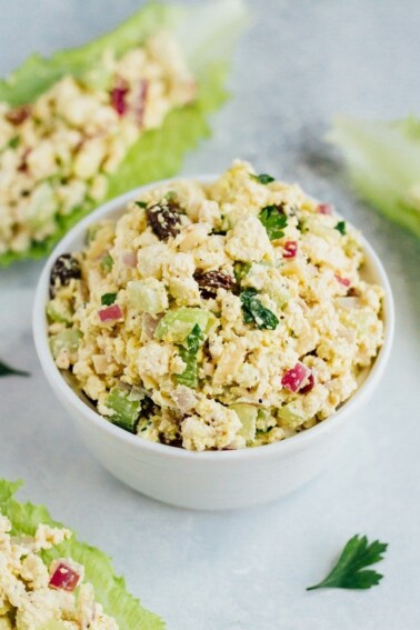 Vegan Chicken Salad made with tofu, celery, almonds and raisins, in a white bowl. Romaine leaves filled with tofu salad sitting around the bowl.