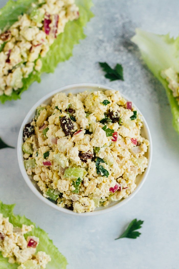 Overhead shot of Vegan Chicken Salad made with tofu, celery, almonds and raisins in a white bowl on white countertop; salad served in romaine.