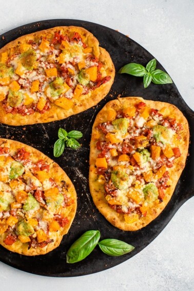 Naan pizza topped with tomatoes, butternut squash, pesto, and cheese on a black baking stone.