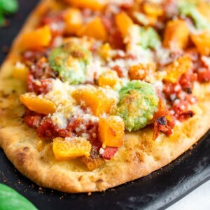 Up close photo of naan pizza topped with tomatoes, butternut squash, pesto, and cheese on a black baking stone.