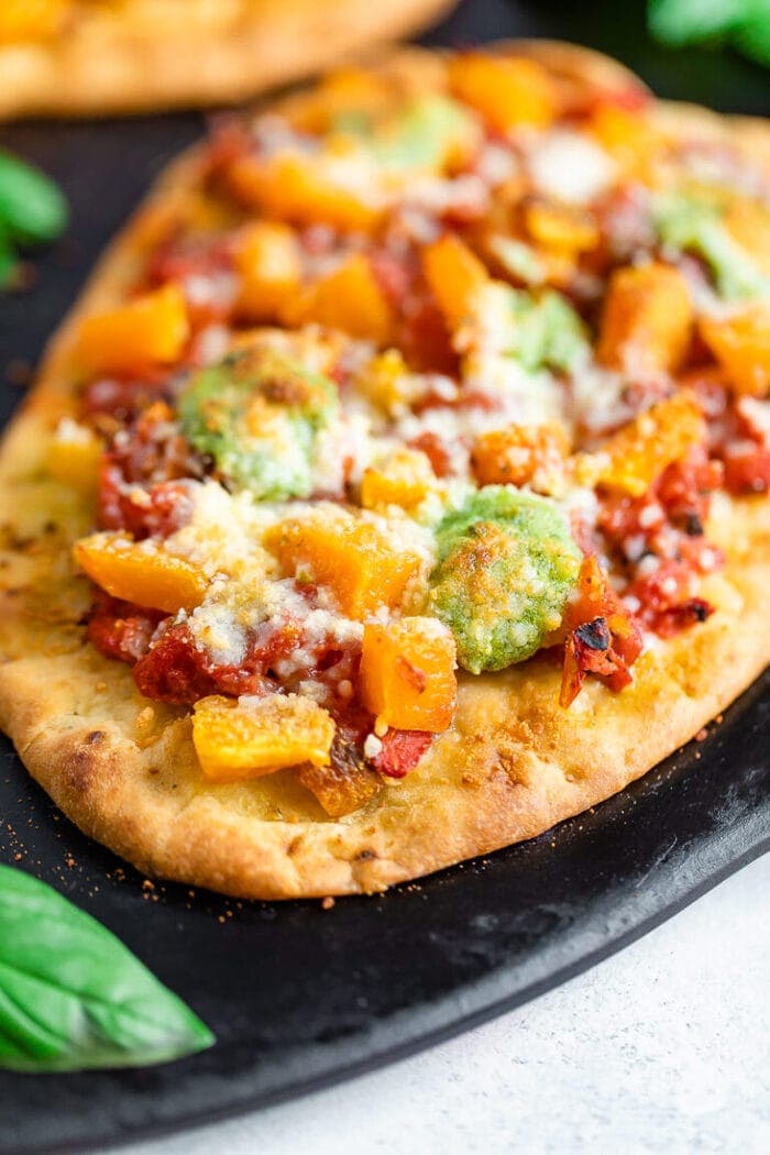 Up close photo of naan pizza topped with tomatoes, butternut squash, pesto, and cheese on a black baking stone.