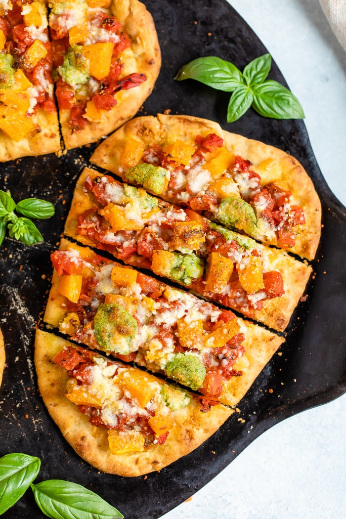 Naan pizza with butternut squash and pesto, sliced into rectangles. Fresh basil is on the baking stone.