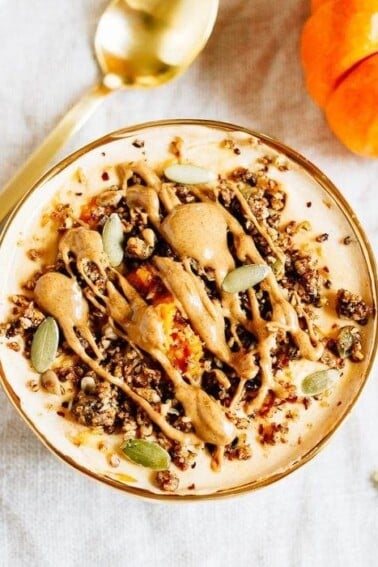 A top down view of a bowl filled with yogurt and pumpkin and topped with seeds, granola and almond butter. Spoon and pumpkin off to the side.