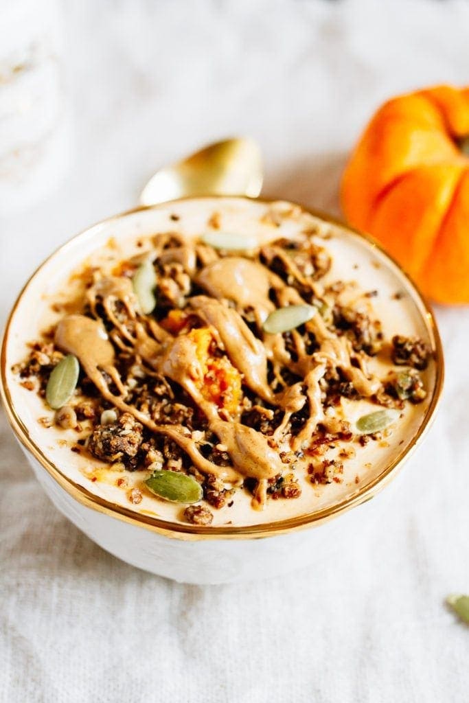 Protein Packed Pumpkin Pie Yogurt Bowl topped with pumpkin, granola, pumpkin seeds, and peanut butter. A mini pumpkin and spoon to the side.