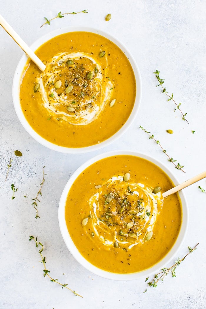 Two bowls of healthy vegan curried pumpkin soup topped with pumpkin seeds and herbs. Sprigs of herbs are around the bowls.