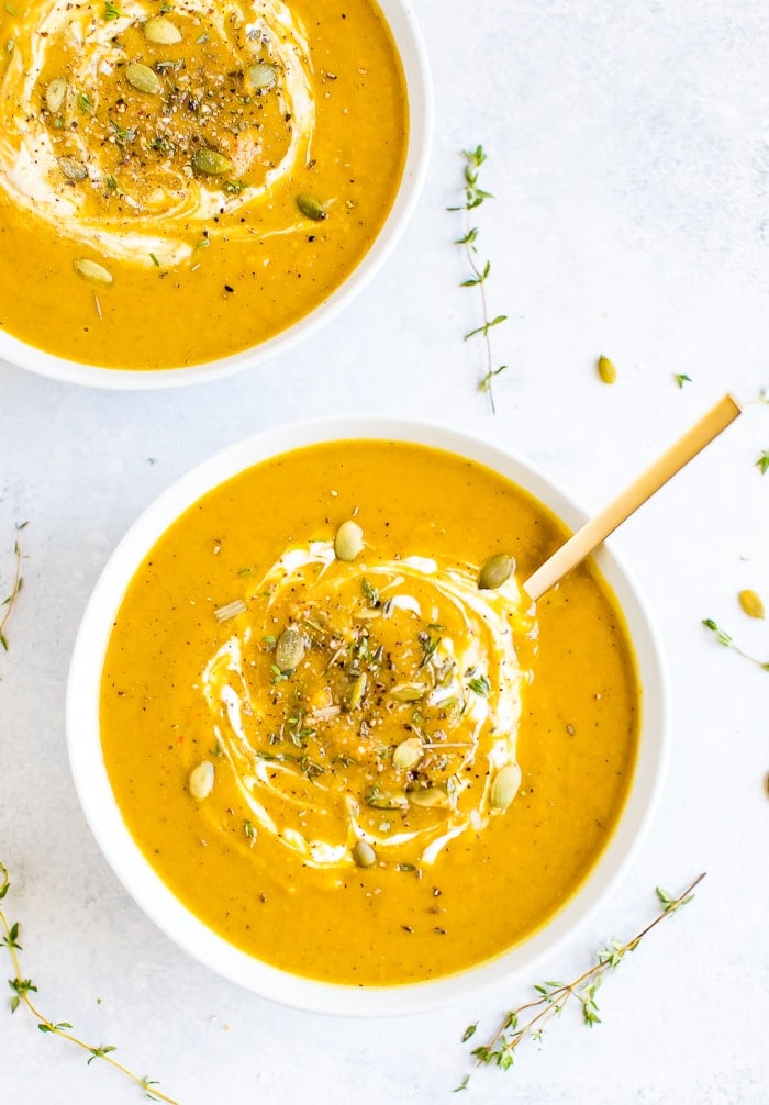 Two bowls of healthy vegan curried pumpkin soup topped with pumpkin seeds and herbs. Sprigs of herbs are around the bowls.