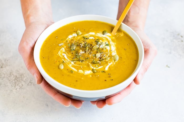 Hands holding a bowl of healthy vegan curried pumpkin soup topped with pumpkin seeds and fresh thyme.