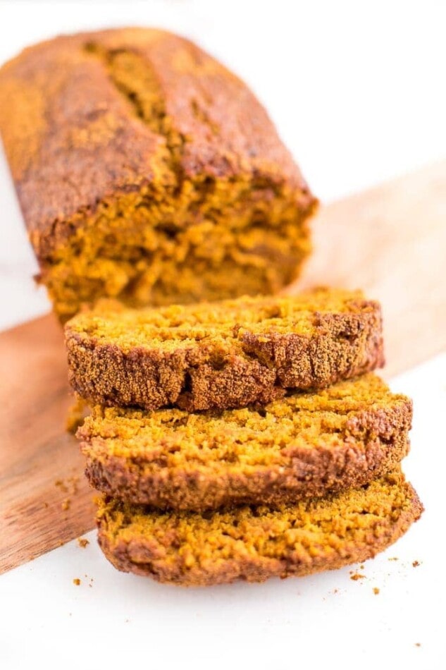 Three slices of low sugar pumpkin bread in the foreground, the remainder of the loaf in the background, all sitting on a cutting board.